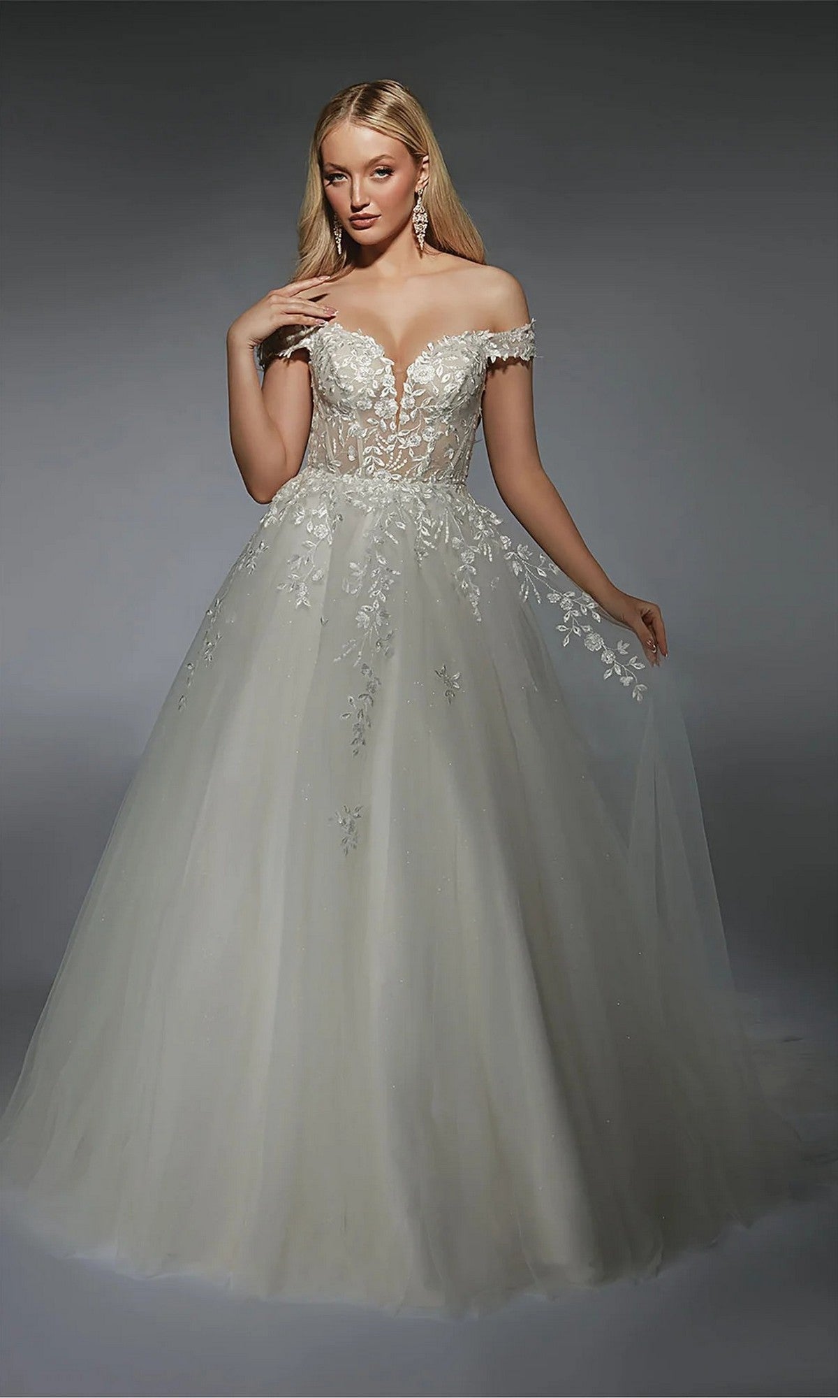 A woman poses against a plain gray background wearing the Alyce 7129, a long ivory dress with an off-the-shoulder lace corset bodice and tulle skirt.